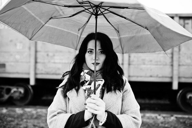 Menina morena de casaco cinza com guarda-chuva vermelho na estação ferroviária