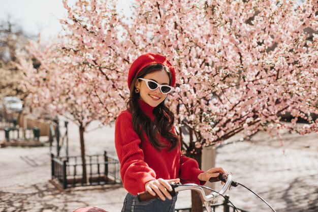 Menina morena de chapéu vermelho e blusa posando no contexto de sakura. Mulher encantadora com óculos de sol elegantes sorrindo e andando de bicicleta