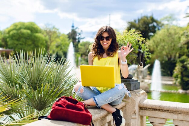 Menina morena de óculos escuros está sentada em cima do muro no parque. Ela segura o laptop amarelo e sorrindo para a câmera.