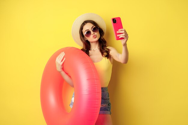 Menina morena elegante de férias, tomando selfie com anel de natação, indo à praia, nadando no mar nas férias de verão, em pé sobre fundo amarelo