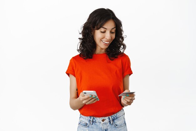 Menina morena elegante faz compras on-line com smartphone e cartão de crédito sorrindo feliz em pé sobre fundo branco