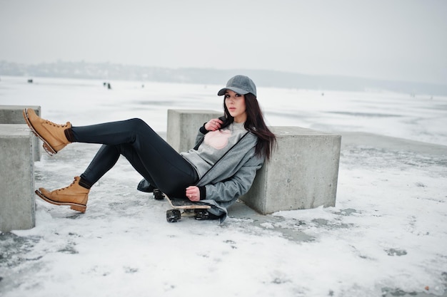 Menina morena elegante no estilo de rua casual de boné cinza com prancha de skate no dia de inverno