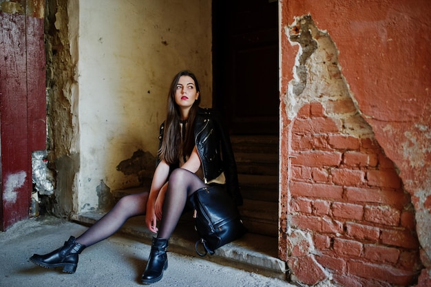 Menina morena elegante usa jaqueta de couro e shorts com mochila sentada em escadas velhas contra a parede laranja
