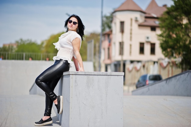 Menina morena em calças de couro femininas e óculos de sol de blusa branca posou contra o skate park