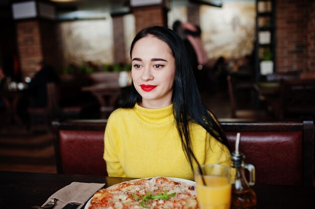 Menina morena engraçada na camisola amarela comendo pizza no restaurante