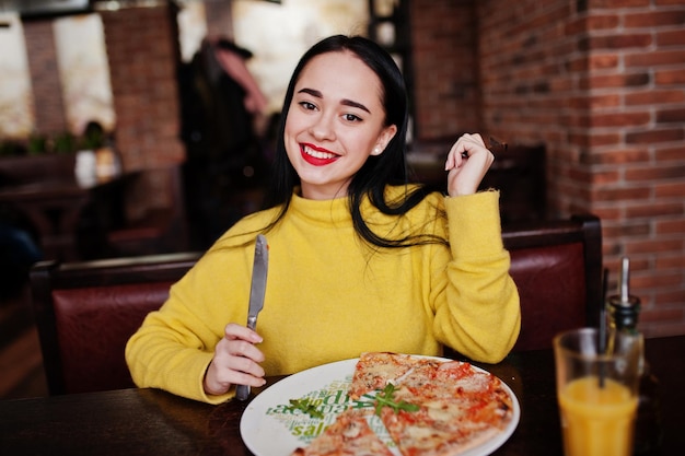 Menina morena engraçada na camisola amarela comendo pizza no restaurante