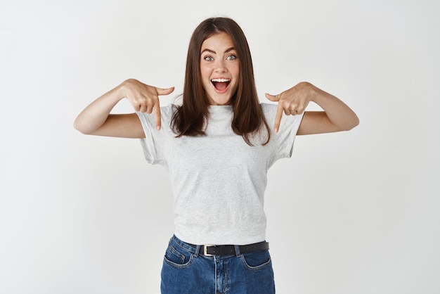 Foto grátis menina morena espantada apontando os dedos para baixo em seu logotipo em pé animado em fundo branco