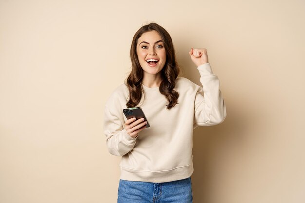 Menina morena feliz segurando o smartphone e torcendo, vencendo, comemorando a vitória no aplicativo para celular, de pé sobre fundo bege.