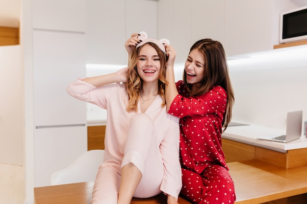 Menina morena glamorosa brincando com a irmã antes do café da manhã no fim de semana. foto interna de duas lindas senhoras sorridentes de pijama.