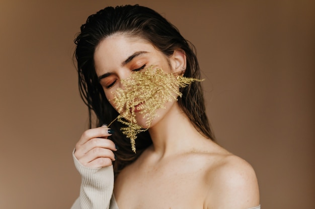 Menina morena graciosa posando com folha verde. mulher caucasiana feliz em pé na parede marrom com a planta.