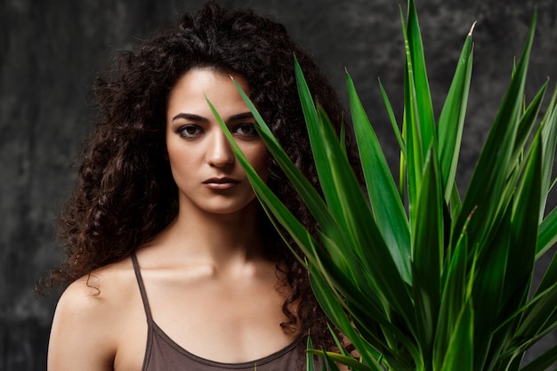 Foto grátis menina morena linda jovem em plantas tropicais ao longo da parede cinza