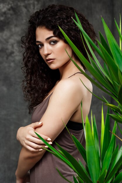 Menina morena linda jovem em plantas tropicais ao longo da parede cinza