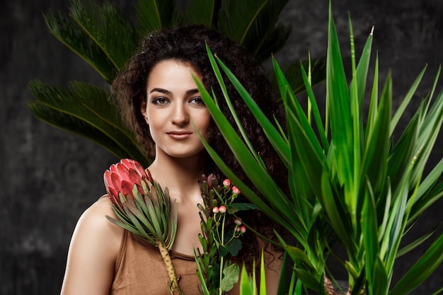 Menina morena linda jovem em plantas tropicais ao longo da parede cinza