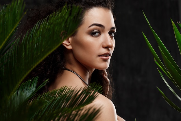 Foto grátis menina morena linda jovem em plantas tropicais ao longo da parede cinza