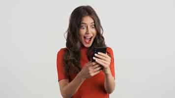 Foto grátis menina morena muito alegre no top vermelho felizmente usando smartphone sobre fundo branco uau expressão
