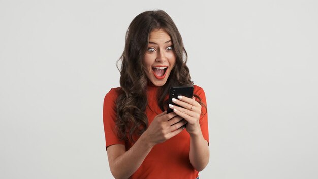 Menina morena muito alegre no top vermelho felizmente usando smartphone sobre fundo branco Uau expressão