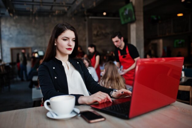 Menina morena sentada no café com uma xícara de cappuccino trabalhando com laptop vermelho