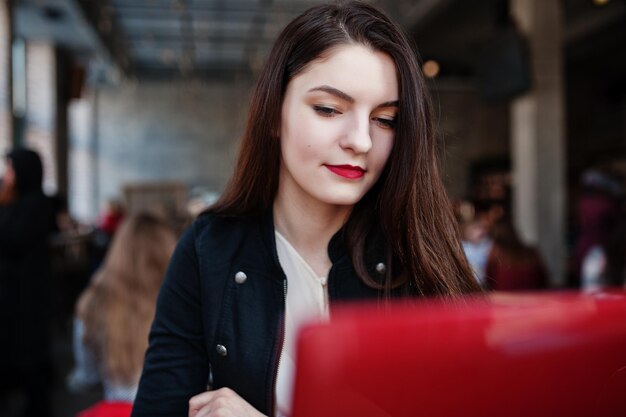 Menina morena sentada no café e trabalhando com laptop vermelho