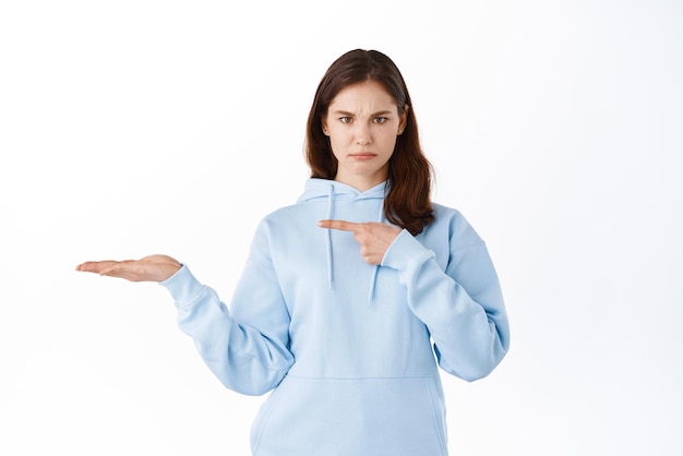 Menina morena triste e irritada apontando para a palma da mão segurando copyspace mostrando o item em exibição na mão em pé contra o fundo branco