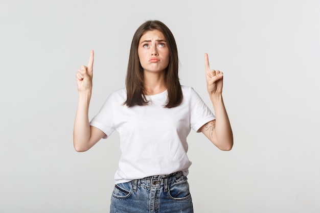 Menina morena triste e sombria de mau humor, apontando e olhando para cima com pesar.