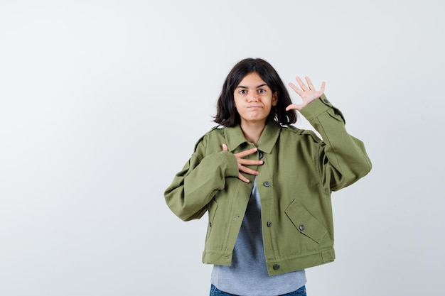 Menina mostrando a palma da mão enquanto segura a mão no peito no casaco, camiseta, jeans e parece confiante, vista frontal.