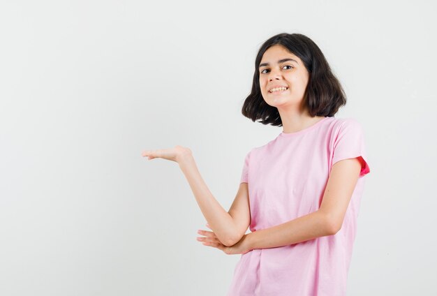 Menina mostrando algo ou dando boas-vindas em t-shirt rosa e olhando alegre, vista frontal.