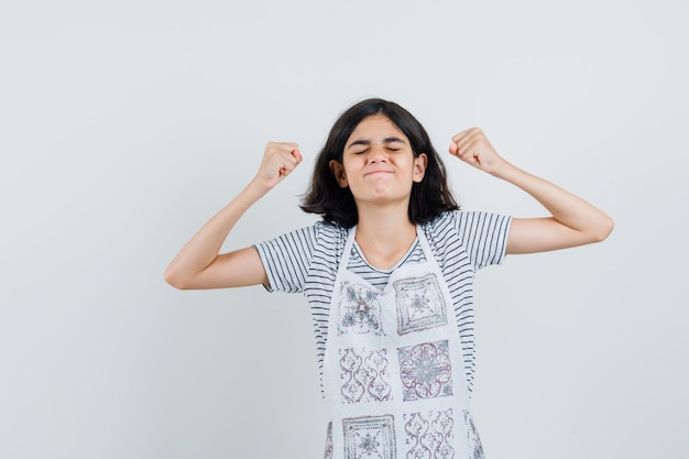 Menina mostrando gesto de vencedor com camiseta, avental e parecendo feliz