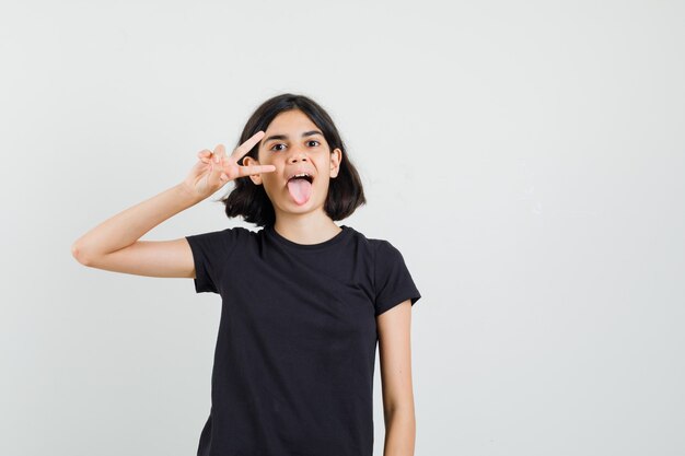 Menina mostrando gesto de vitória em t-shirt preta e parecendo confiante. vista frontal.