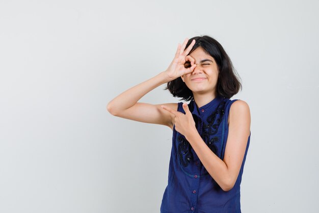 Menina mostrando sinal de ok no olho, apontando para longe com uma blusa azul e parecendo brincalhona