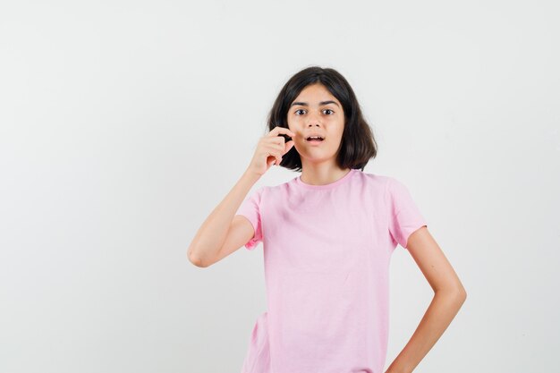 Menina mostrando sinal de tamanho pequeno em t-shirt rosa e olhando surpresa, vista frontal.