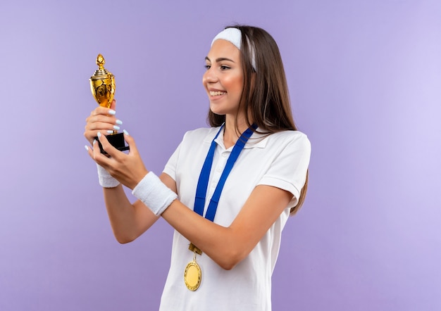 Foto grátis menina muito esportiva sorridente usando bandana, pulseira e medalha segurando e olhando para o copo isolado no espaço roxo