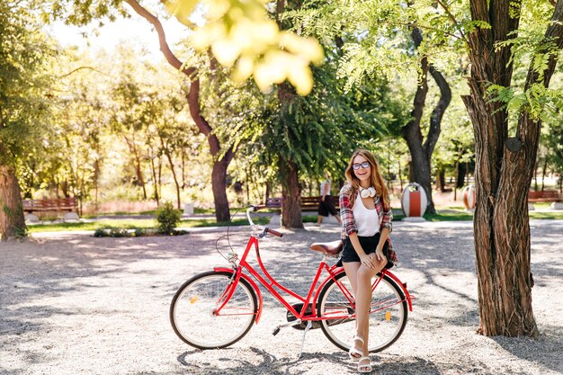 Menina muito magra sentada na bicicleta vermelha. Mulher elegante de Jocund, aproveitando o fim de semana ativo.