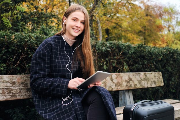 Menina muito sorridente com mala felizmente usando tablet descansando no banco no parque da cidade