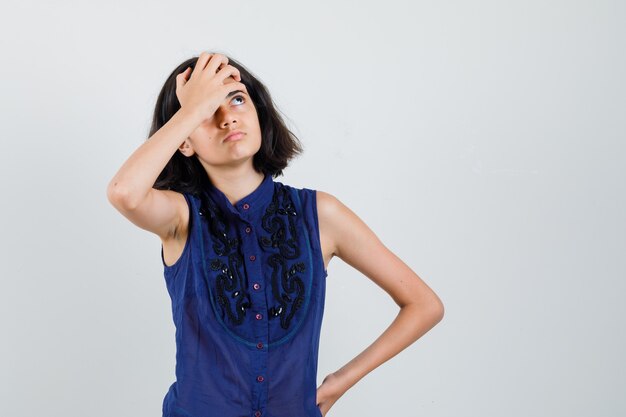 Menina na blusa azul, segurando a mão na testa e olhando com foco, vista frontal.