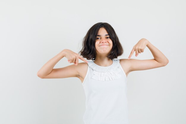 Menina na blusa branca, apontando para si mesma e orgulhosa, vista frontal.