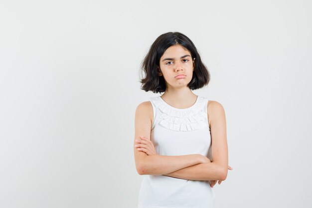 Menina na blusa branca em pé com os braços cruzados e olhando chateada, vista frontal.