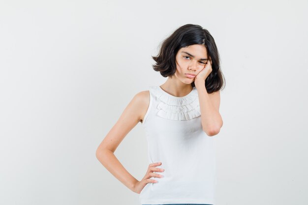 Menina na blusa branca, inclinando a bochecha na palma da mão e parecendo cansada, vista frontal.