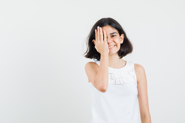 Menina na blusa branca, segurando a mão no olho e olhando alegre, vista frontal.