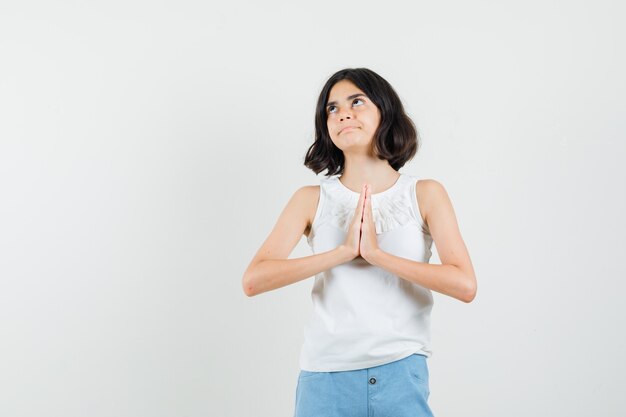 Menina na blusa branca, shorts de mãos dadas em gesto de oração e olhando esperançosa, vista frontal.
