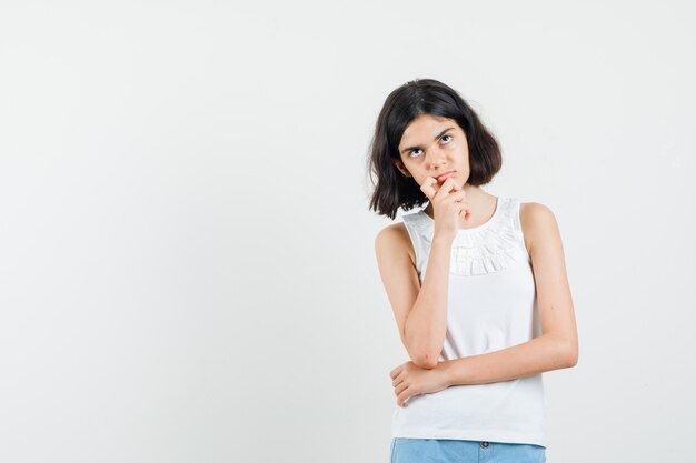 Menina na blusa branca, shorts olhando para cima e parecendo pensativo, vista frontal.