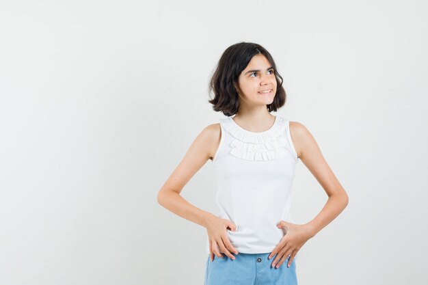 Menina na blusa branca, shorts olhando para longe e parecendo alegre, vista frontal.