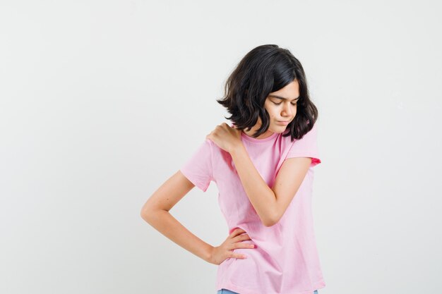 Menina na camiseta rosa com dor no ombro e parecendo cansada, vista frontal.