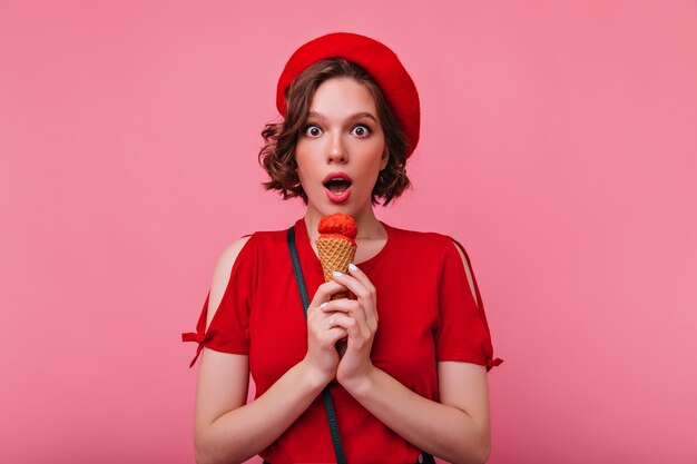 Menina na moda tomando sorvete e expressando espanto. Foto interna de adorável francesa na boina vermelha elegante.
