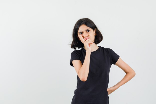 Menina na t-shirt preta em pé em pose de pensamento e olhando sério, vista frontal.