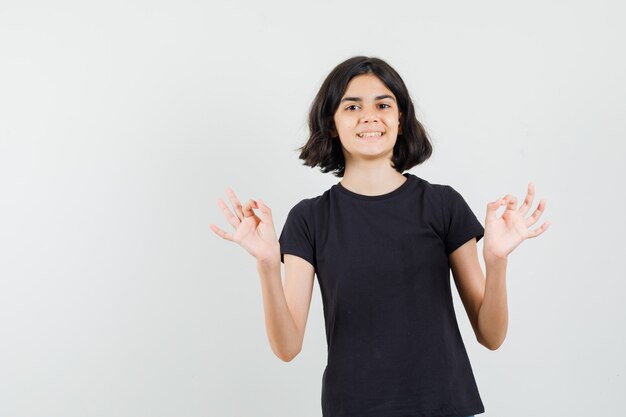Menina na t-shirt preta, mostrando o sinal de ok e olhando alegre, vista frontal.