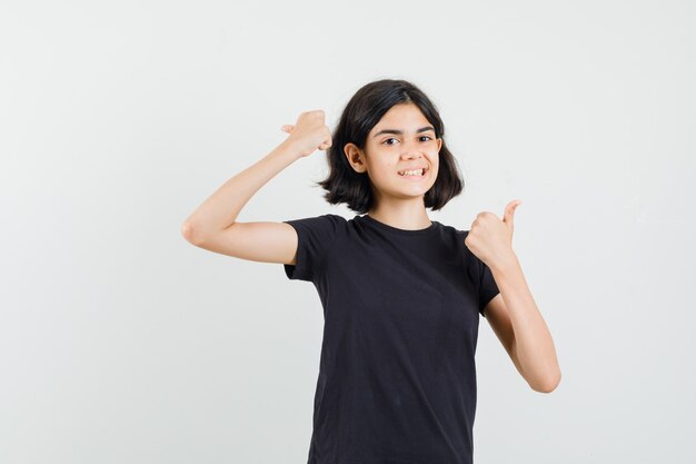 Menina na t-shirt preta, mostrando os polegares duplos e olhando alegre, vista frontal.