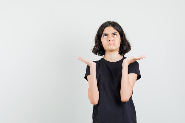 Menina na t-shirt preta, mostrando um gesto de impotência e parecendo confusa, vista frontal.