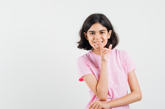 Menina na t-shirt rosa em pé em pose de pensamento e olhando alegre, vista frontal.