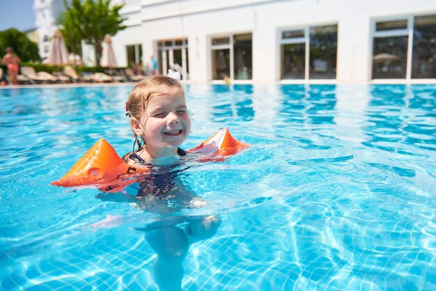 Menina nadando na piscina em braçadeiras em um dia quente de verão. Férias em família em resort tropical