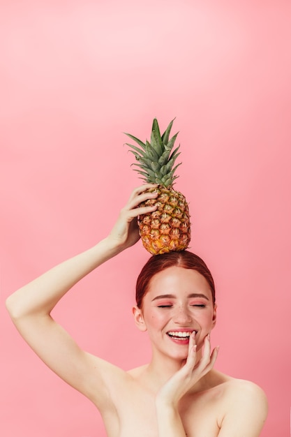 Menina nua com abacaxi, sorrindo com os olhos fechados. Foto de estúdio de gengibre jovem segurando frutas exóticas.